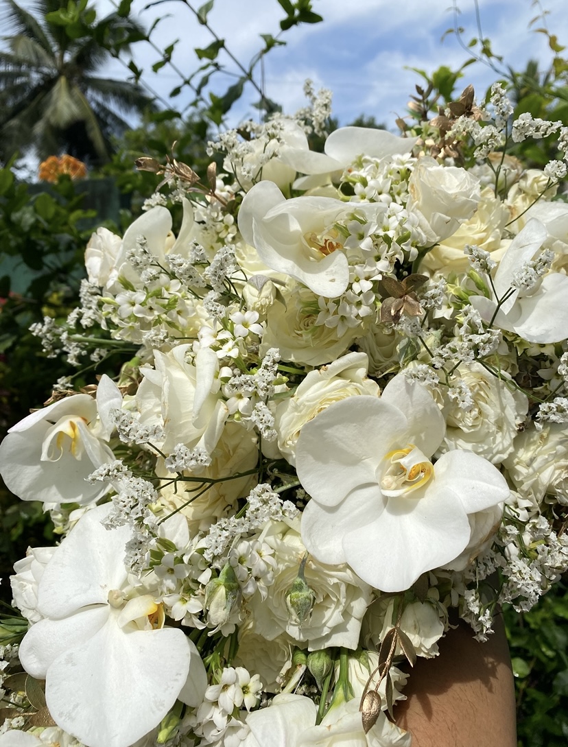 A fresh white wedding bouquet of roses and orchids, held up against palm trees and blue sky